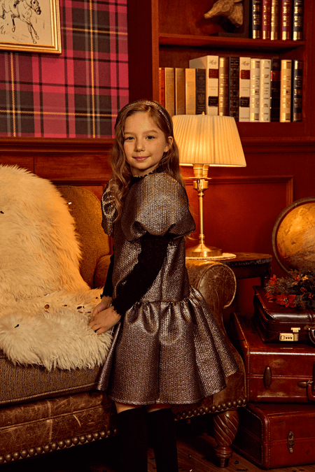 Young girl in Selena Jacquard Balloon Dress, light gold, standing by a cozy library setting with elegant decor and plush armchair.