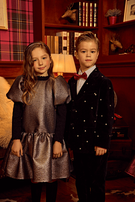 Two children in formal attire, with the girl wearing a light gold Selena Jacquard Balloon Dress, standing in a warmly lit room with a classic decor.