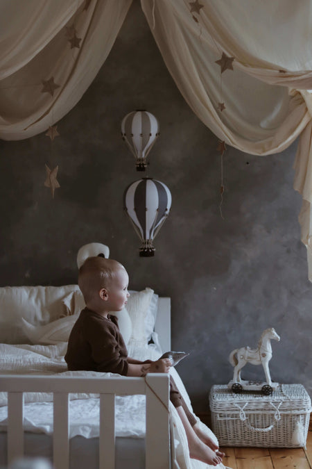 Child in nursery with white and dark grey hot air balloon decor, creating a whimsical atmosphere with star canopy and toy horse.