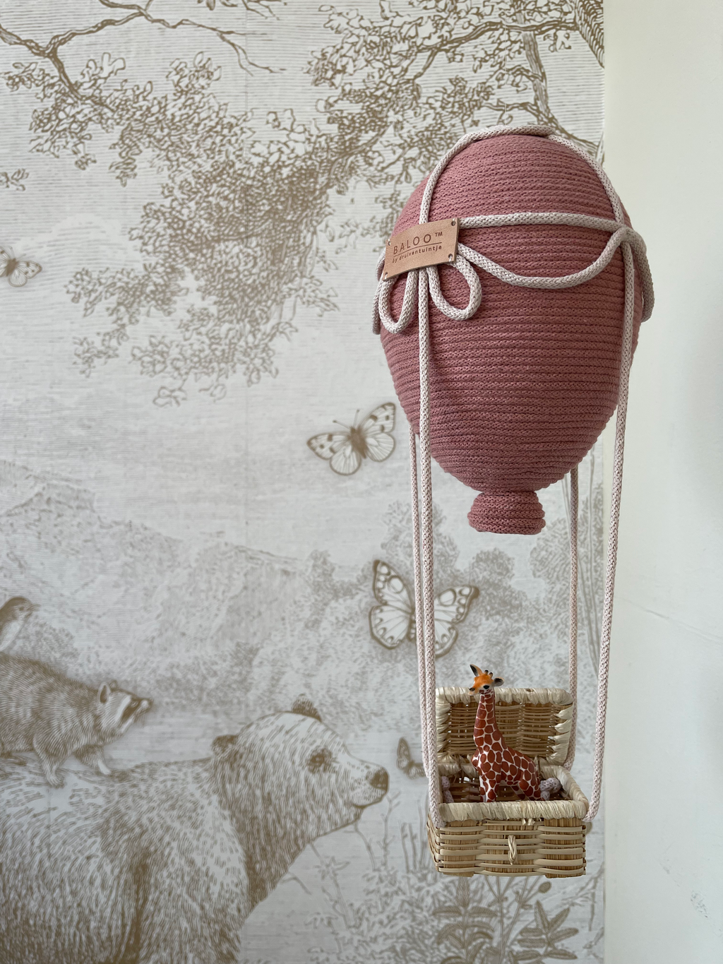 Blush Baloo hot air balloon decoration for baby nursery with giraffe in basket, handcrafted detail, against animal-themed wallpaper.